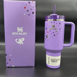 Puede incluir: Un vaso Stanley Quencher H2.0 morado con asa, pajita y tapa, junto a una caja a juego. El vaso y la caja est&aacute;n decorados con gr&aacute;ficos de estrellas y mariposas. El vaso contiene 1,18L.