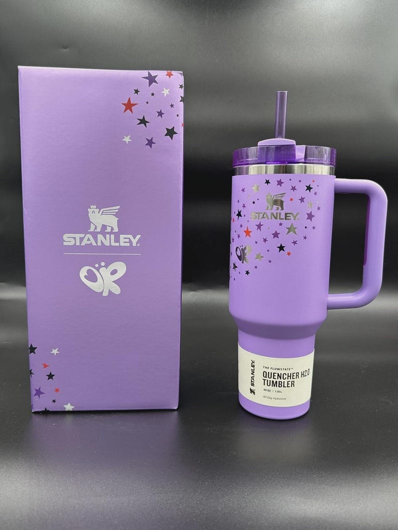 Puede incluir: Un vaso Stanley Quencher H2.0 morado con asa, pajita y tapa, junto a una caja a juego. El vaso y la caja est&aacute;n decorados con gr&aacute;ficos de estrellas y mariposas. El vaso contiene 1,18L.