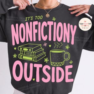 May include: A charcoal grey crewneck sweatshirt featuring the text IT'S TOO NONFICTIONY OUTSIDE in large pink letters. The design includes a stack of three books and a steaming mug in lime green, accented with small green and pink floral motifs.