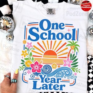 May include: White t-shirt with a colourful graphic. The design features the text "One School Year Later" in blue, with a sunset, palm trees, and floral elements in orange, pink, and green. The shirt has a crew neck and a Comfort Colors tag.
