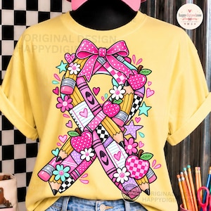 May include: A pale yellow t-shirt featuring a large graphic of a ribbon shape constructed from pink and yellow pencils, lined paper, and checkered patterns. The design includes pink bows, small white flowers, teal stars, and pink polka-dot apples.
