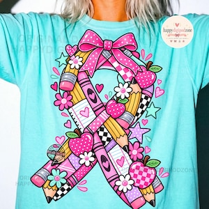 May include: A light turquoise t-shirt featuring a large awareness ribbon graphic composed of yellow pencils, pink crayons, lined paper, and red apples. The design includes a pink polka-dot bow, small flowers, and stars. Text reads "happydigipodzone".