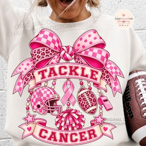 May include: A white sweatshirt featuring a large pink graphic of a bow, a football helmet, a football, a whistle, and a pom-pom. The text TACKLE CANCER is printed in pink block letters. A pink ribbon symbol is centered between the sports items.