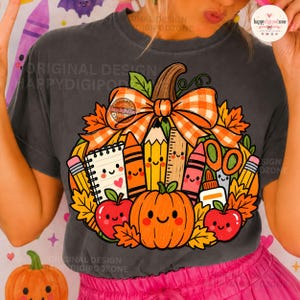 Peut inclure: Un t-shirt gris anthracite orné d'un grand motif représentant une citrouille décorée de fournitures scolaires, dont un carnet, des crayons, des craies, une règle et des ciseaux, surmontée d'un nœud à carreaux orange et de feuilles d'automne.