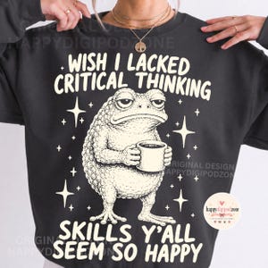 Puede incluir: Una sudadera negra de cuello redondo con un gráfico de color crema de un sapo sosteniendo una taza. El texto dice WISH I LACKED CRITICAL THINKING SKILLS Y'ALL SEEM SO HAPPY. El diseño incluye pequeñas estrellas que rodean al sapo al frente.