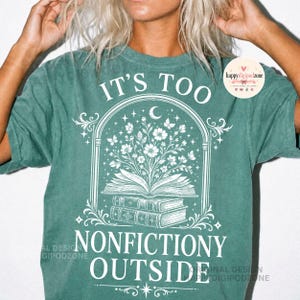 May include: A teal short-sleeve t-shirt featuring a white graphic of an open book with flowers blooming from the pages, stacked on two other books. The text reads IT'S TOO NONFICTIONY OUTSIDE, framed by an arched design with stars and a crescent moon.