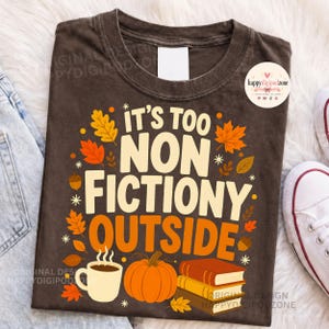 May include: A dark brown t-shirt featuring the text IT'S TOO NON FICTIONY OUTSIDE in cream and orange lettering. The design includes orange and yellow autumn leaves, a white coffee mug with steam, an orange pumpkin, and a stack of three books.