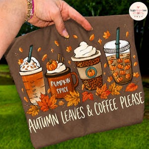 Puede incluir: Una camiseta marrón con un gráfico de cuatro tazas de café con motivos otoñales, incluyendo el texto "pumpkin spice", nata montada y hojas naranjas. El texto en la parte inferior dice "AUTUMN LEAVES & COFFEE PLEASE" en una fuente clara.