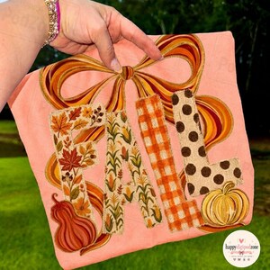May include: A peach-colored t-shirt featuring the word FALL in large letters. The F has a leaf pattern, the A has a corn stalk pattern, the L has an orange gingham check, and the second L has brown polka dots, all topped with a large bow.