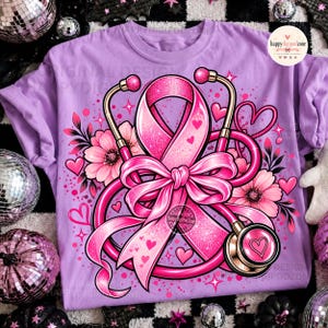 May include: A light purple t-shirt featuring a central graphic of a glittery pink awareness ribbon intertwined with a stethoscope, surrounded by pink flowers, small hearts, and star accents. The design includes a circular logo in the top right corner.