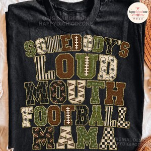 Può includere: Una t-shirt nera con il testo SOMEBODY'S LOUD MOUTH FOOTBALL MAMA in grandi lettere stampatello. Le lettere presentano vari motivi tra cui palloni da football, caschi, strisce e segni di campo in tonalità marrone, verde e bianco.