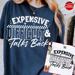 May include: A navy blue t-shirt featuring white text that reads EXPENSIVE DIFFICULT & Talks Back, accented with a checkered pattern and small star icons. A smaller white t-shirt with the same black graphic design is shown in the bottom corner.