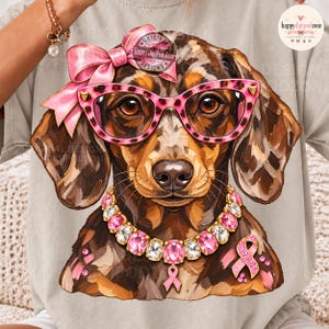 May include: A beige t-shirt featuring a graphic of a brown dachshund wearing pink leopard-print glasses, a large pink bow, and a sparkling pink and white crystal necklace. A pink awareness ribbon is positioned on the dog's chest area.
