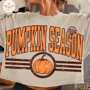 Può includere: Una felpa beige a girocollo con la scritta PUMPKIN SEASON in grandi lettere stampatello color arancione usurato. Sotto il testo, una grafica circolare contiene l'illustrazione di una zucca arancione tra due strisce orizzontali marroni.