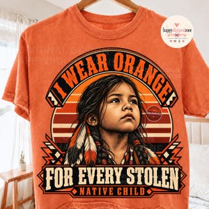 Könnte beinhalten: Ein orangefarbenes T-Shirt mit einer Grafik eines Kindes mit geflochtenem Haar und Federn. Der Text lautet I WEAR ORANGE FOR EVERY STOLEN NATIVE CHILD. Das Design zeigt einen Sonnenuntergangshintergrund mit Streifen und geometrischen Elementen.