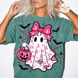 May include: A teal t-shirt featuring a white ghost graphic with a large pink bow, pink breast cancer awareness ribbons, and small hearts. The ghost holds a pink jack-o'-lantern bucket, surrounded by black spiderwebs, spiders, and bats.