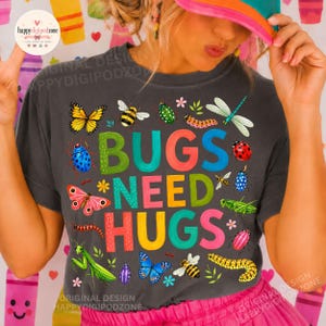 May include: A dark gray t-shirt featuring the text BUGS NEED HUGS in colorful, patterned letters. The design is surrounded by various illustrated insects, including a yellow butterfly, a bee, a ladybug, a dragonfly, and a green caterpillar.