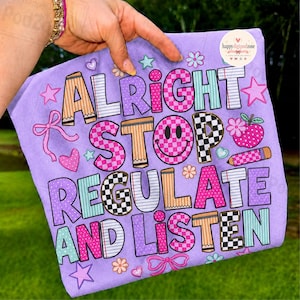 Pode incluir: Uma t-shirt roxo-claro com o texto ALRIGHT STOP REGULATE AND LISTEN em letras de bloco coloridas e estampadas. O design inclui elementos gráficos como estrelas, corações, laços, uma cara sorridente, um lápis e uma flor.