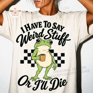 May include: A cream-colored t-shirt featuring a graphic of a green frog holding a brown mug. The text reads I HAVE TO SAY Weird Stuff Or I'll DIE. Behind the frog is a black and white checkered pattern and small yellow star shapes.