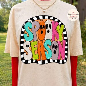 May include: A cream-coloured t-shirt featuring a graphic with the text SPOOKY SEASON in colourful, stylised letters shaped like ghosts. The design is enclosed in a black and white striped border. A gold-toned chain necklace is worn around the neck.