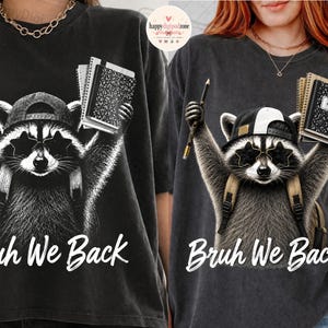 May include: A black t-shirt featuring a graphic of a raccoon wearing a baseball cap and star-shaped sunglasses. The raccoon holds a pencil and a notebook with both arms raised. Below the graphic, the white text reads, Bruh We Back.