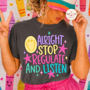 May include: A charcoal gray short-sleeve t-shirt featuring a graphic print with the text "ALRIGHT STOP REGULATE AND LISTEN" in purple, yellow, pink, and teal. The design includes a yellow smiley face and several pink and teal stars.