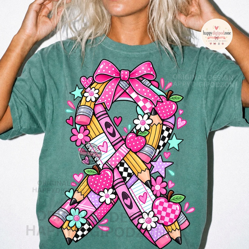 May include: A muted green t-shirt featuring a large graphic of a awareness ribbon constructed from yellow pencils, pink crayons, and a pink bow. The design includes small pink apples, white flowers, and teal stars scattered around the central ribbon motif.