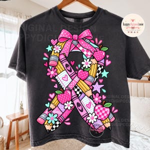 May include: A charcoal gray t-shirt featuring a large graphic of a awareness ribbon composed of yellow pencils, pink crayons, and checkered patterns. The design includes pink bows, red and pink apples, white flowers, and small hearts and stars.