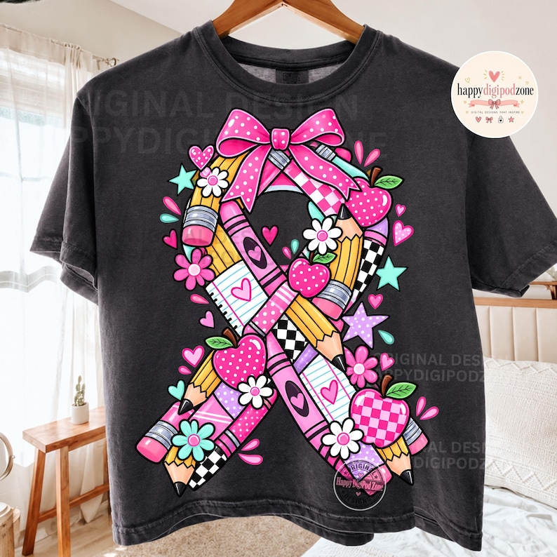 May include: A charcoal gray t-shirt featuring a large graphic of a awareness ribbon composed of yellow pencils, pink crayons, and checkered patterns. The design includes pink bows, red and pink apples, white flowers, and small hearts and stars.