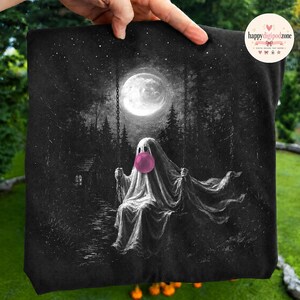 May include: A black fabric item featuring a white graphic illustration of a ghost sitting on a swing under a full moon in a forest. The ghost is blowing a bright pink bubble with bubblegum. The scene includes a small cabin and starry night sky.