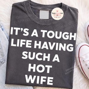 Puede incluir: Una camiseta de color gris carbón con cuello redondo que presenta el texto blanco "IT'S A TOUGH LIFE HAVING SUCH A HOT WIFE" impreso en una fuente negrita sans-serif en el pecho. La prenda tiene un cuello acanalado y un acabado desgastado.