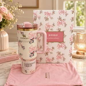 May include: A cream-colored Stanley Quencher H2.0 tumbler featuring a pink floral and ribbon pattern, a matching pink handle, and a clear lid. It is accompanied by a rectangular box with the same pattern and a pink fabric drawstring storage bag.