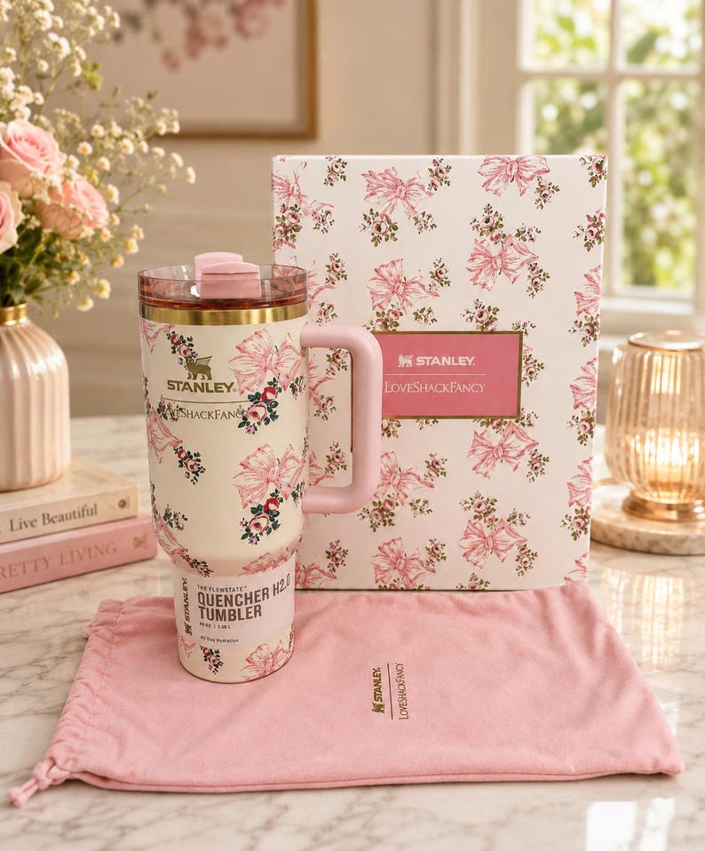 May include: A cream-colored Stanley Quencher H2.0 tumbler featuring a pink floral and ribbon pattern, a matching pink handle, and a clear lid. It is accompanied by a rectangular box with the same pattern and a pink fabric drawstring storage bag.