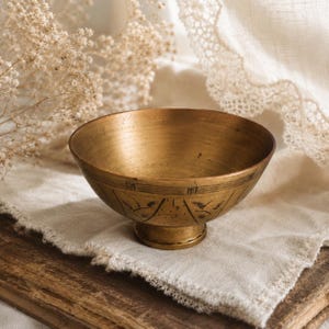 May include: A brass bowl with a wide, open top and a pedestal base. The exterior surface features etched black line details, including horizontal bands and repeating geometric patterns with small leaf-like motifs encircling the lower section.