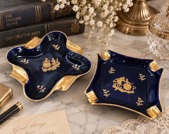 May include: Two square-shaped, deep blue ceramic trays with scalloped edges and gold-toned rims. Each tray features a central gold-colored illustration of a couple in period clothing, surrounded by small gold floral accents on the interior surface.