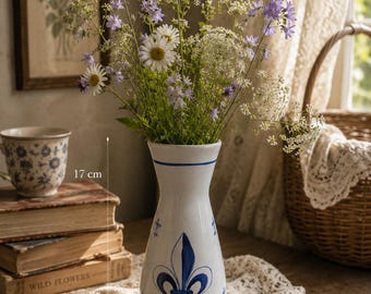 May include: A white ceramic vase with a blue fleur-de-lis pattern and a blue horizontal line near the rim. The vase stands 17 cm tall and is filled with a bouquet of white daisies, small purple bell-shaped flowers, and delicate white lace-like wildflowers.