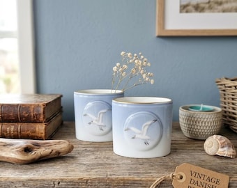 May include: Two light blue ceramic mugs with a seagull design, set on a wooden shelf. The mugs have a gold rim and are part of a vintage Danish B&G set. A sprig of white flowers is in one mug.