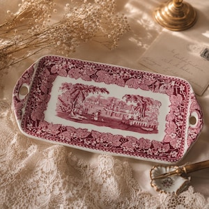 Puede incluir: Una bandeja de servicio rectangular de cerámica con un patrón de transferencia en color rojo oscuro. El centro representa un paisaje con edificios y árboles, mientras que el borde muestra un denso motivo de hojas de parra y dos asas.