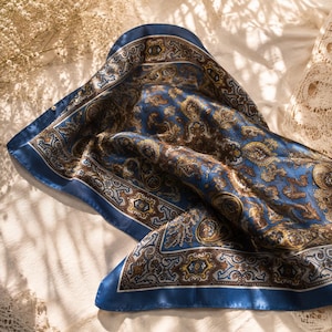 May include: A square silk scarf featuring a deep blue base color with an intricate paisley pattern in shades of brown, gold, and white. The scarf has a wide, solid blue border and a detailed central design with repeating ornate motifs.