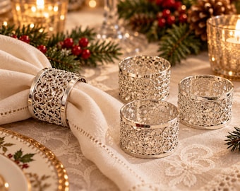 May include: A set of four silver-toned metal napkin rings featuring a circular filigree pattern. One ring holds a folded cream-colored fabric napkin, while three identical rings are arranged nearby on a table surface.