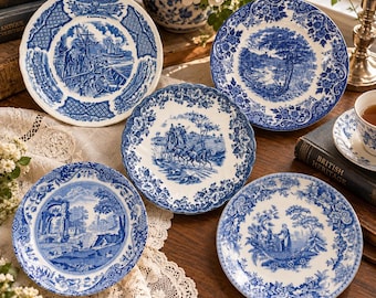 May include: A collection of five round ceramic plates featuring blue and white transferware patterns. Each plate displays detailed historical scenes, including a ship, a horse-drawn carriage, and pastoral landscapes, all framed by intricate floral borders.