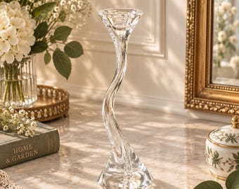 May include: A clear glass candle holder featuring a tall, slender, and wavy stem that rises from a wide, circular base to a flared, circular top opening. The glass is transparent and smooth, reflecting light along its curved, organic silhouette.