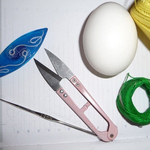 May include: A collection of craft supplies including a blue decorative piece, pink handled scissors, a crochet hook, a white egg, a ball of green thread, and a spool of yellow thread. The items are arranged on a lined surface.