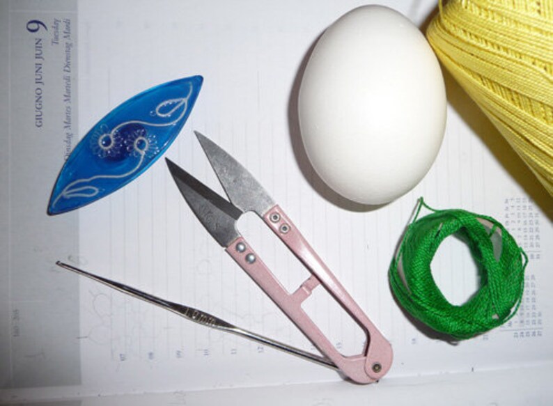 May include: A collection of craft supplies including a blue decorative piece, pink handled scissors, a crochet hook, a white egg, a ball of green thread, and a spool of yellow thread. The items are arranged on a lined surface.