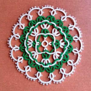 Puede incluir: Un encaje de ganchillo blanco y verde con un diseño circular. El encaje tiene un círculo central con cuatro círculos más pequeños alrededor, todos conectados por una serie de bucles.