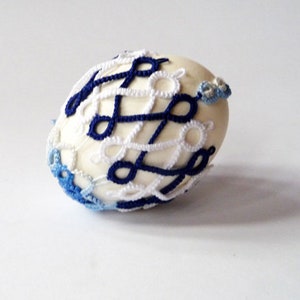 May include: A decorative egg with a cream-colored base, intricately adorned with a crocheted pattern in navy blue and white. The egg features a delicate, openwork design, with accents of light blue crochet at the ends.