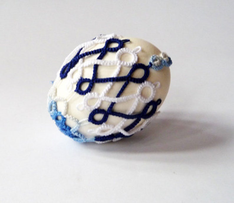 May include: A decorative egg with a cream-colored base, intricately adorned with a crocheted pattern in navy blue and white. The egg features a delicate, openwork design, with accents of light blue crochet at the ends.