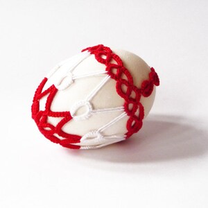 May include: A cream-colored egg is adorned with a red and white crocheted design. The red crochet forms a band around the egg, while the white crochet creates a web-like pattern. The egg is set against a white background.