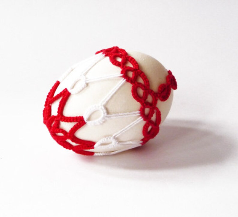 May include: A cream-colored egg is adorned with a red and white crocheted design. The red crochet forms a band around the egg, while the white crochet creates a web-like pattern. The egg is set against a white background.