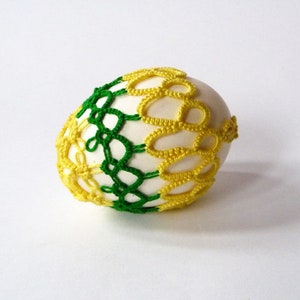 May include: A decorative egg with a white base, adorned with a crocheted overlay. The crochet design features a combination of green and yellow threads, creating a patterned, textured appearance. The egg is a craft item, likely for Easter or home decor.